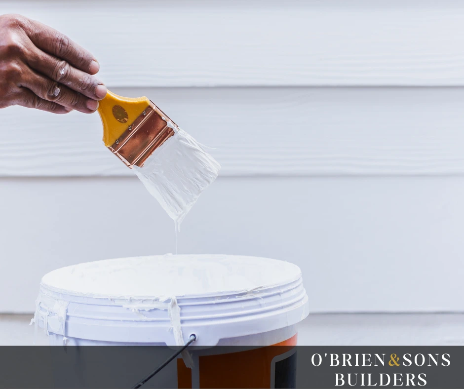 Hand dipping a paintbrush into a bucket of high-quality white paint in front of a clean exterior wall in Tarzana, CA.